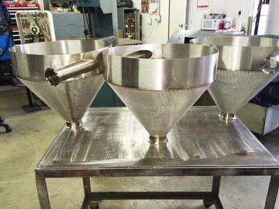 Three fabricated stainless steel hoppers with perforated cones and side inlets on a stainless workbench