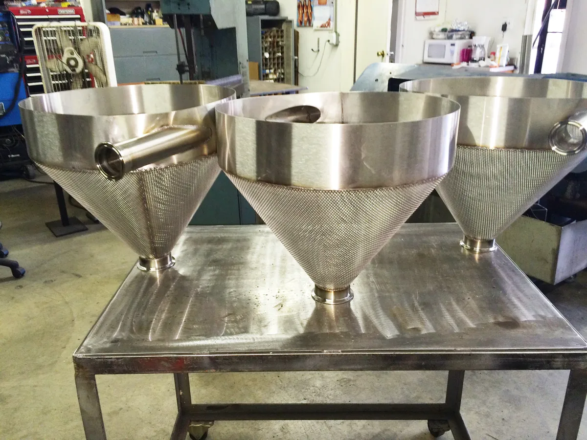 Three fabricated stainless steel hoppers with perforated cones and side inlets on a stainless workbench