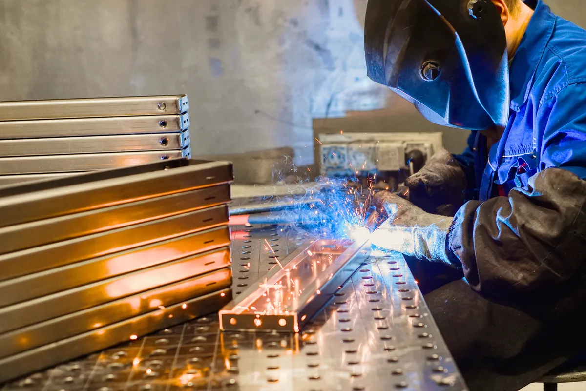 Welder joining stainless steel profiles on a fixture table beside a stack of finished parts
